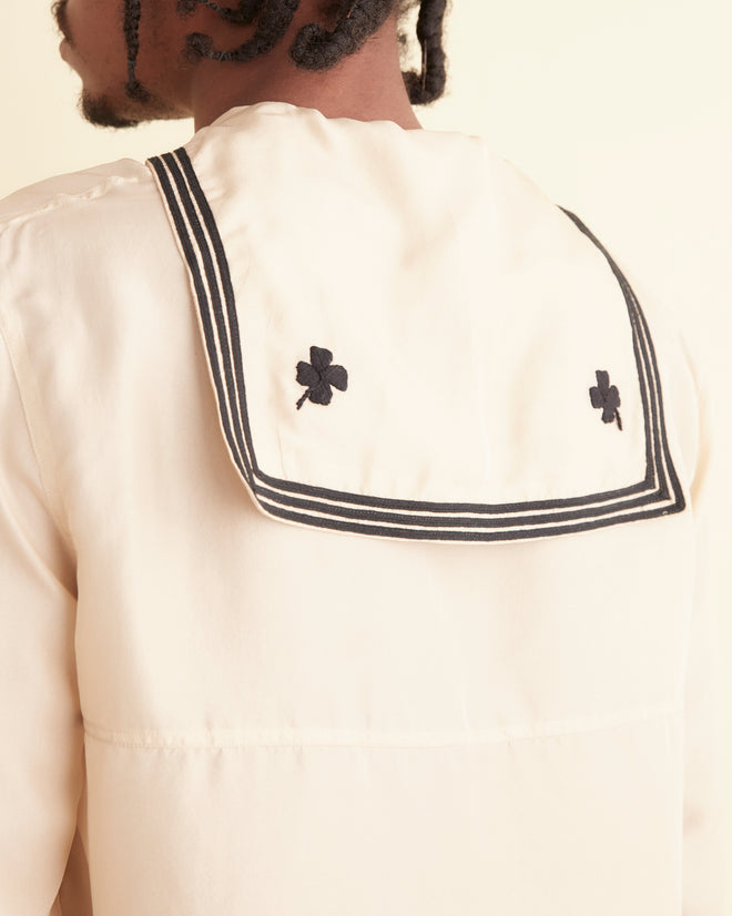 A person stands against an off-white wall wearing an ecru-colored Cricket Shirt with a sailor collar and black pants. They have braided hair and are looking off to the side.