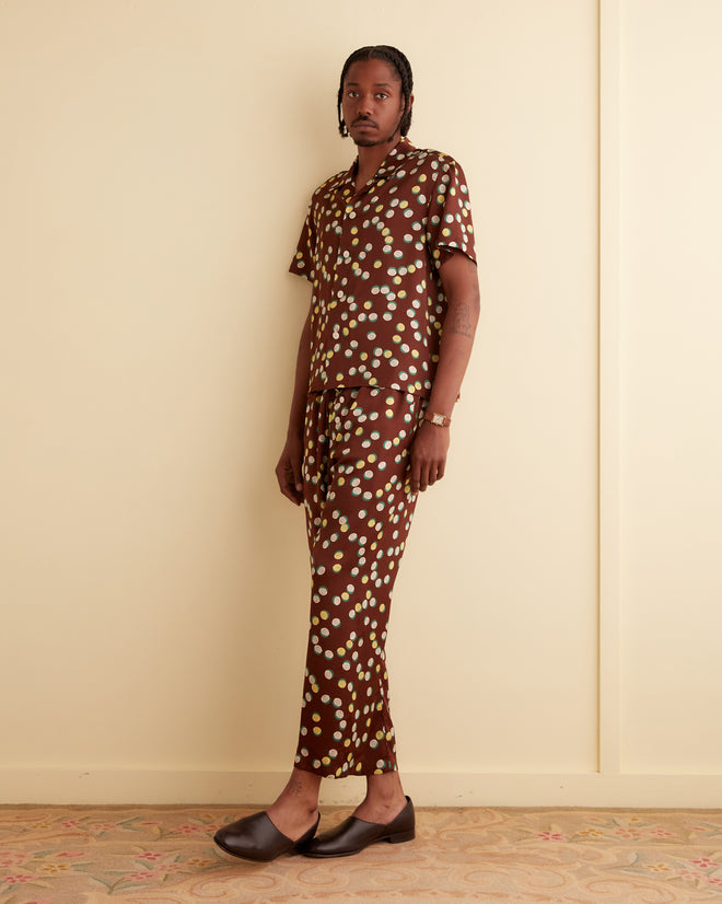 A person stands against a beige wall wearing the Bubble Dot Short Sleeve Shirt, which features a brown outfit with white polka dots reminiscent of the 1930s-1940s style. They have braided hair and are wearing dark shoes.