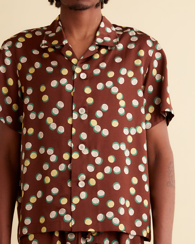 A person stands against a beige wall wearing the Bubble Dot Short Sleeve Shirt, which features a brown outfit with white polka dots reminiscent of the 1930s-1940s style. They have braided hair and are wearing dark shoes.