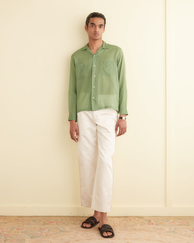 A person wearing a green Voile Long Sleeve Shirt, white pants, sandals, and a wristwatch, standing against an off-white wall on a patterned carpet.