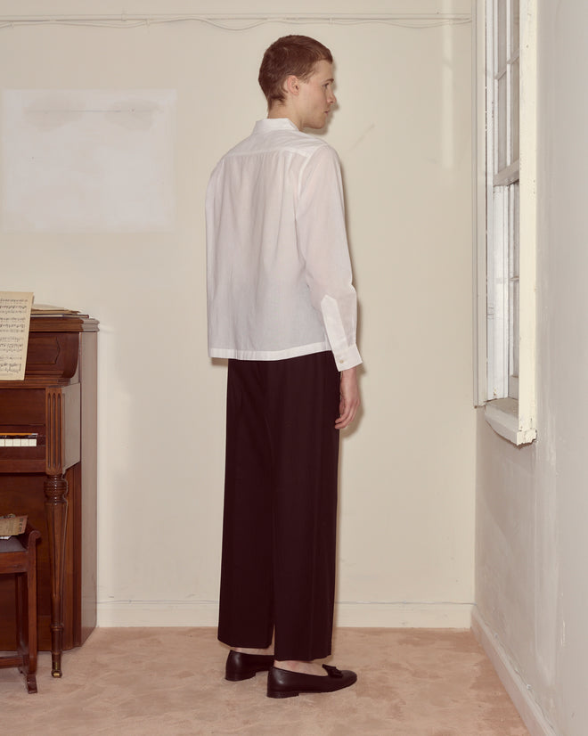 Wearing the Voile Long Sleeve Shirt - White and black pants, a person stands facing a wall beside a piano and window in a beige room, highlighting the shirt’s boxy silhouette.