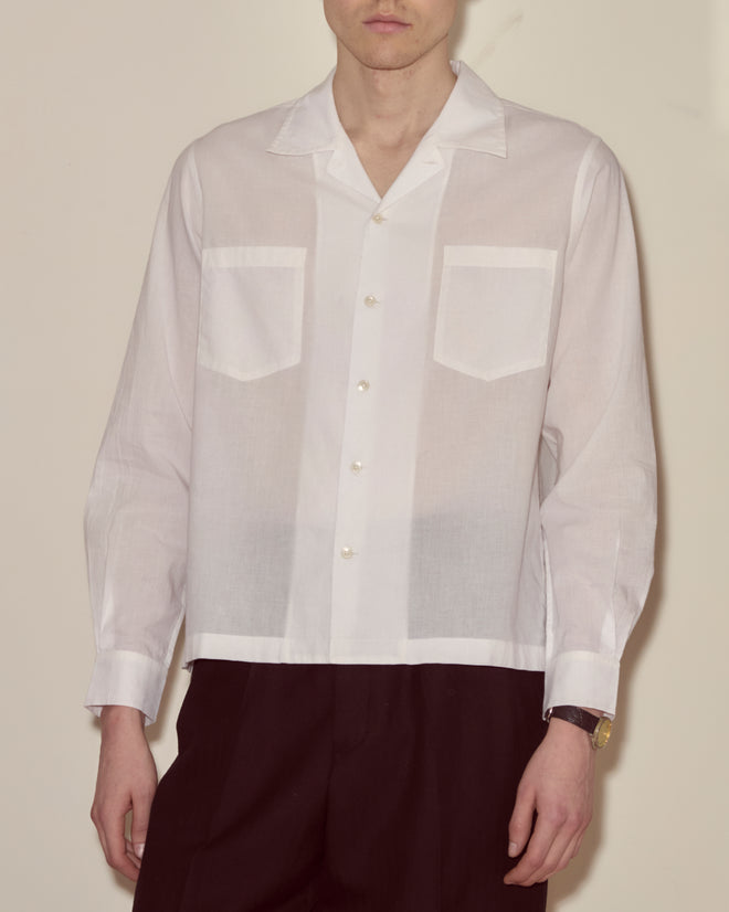 A man models the Voile Long Sleeve Shirt - White, crafted from sheer 100% cotton in a boxy fit, paired with black pants against a plain background.