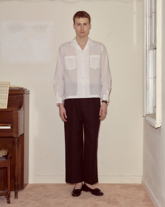 A man wearing the Voile Long Sleeve Shirt - White and black pants stands indoors by a piano and window, looking at the camera.