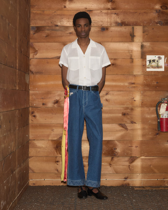 A person in a white short-sleeve shirt and Embroidered Denim Knolly Brook Trousers stands against a wooden wall with a fire extinguisher nearby.