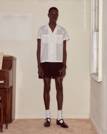 Wearing the Voile Short Sleeve Shirt - White and black shorts, a person stands indoors by a window next to a piano.