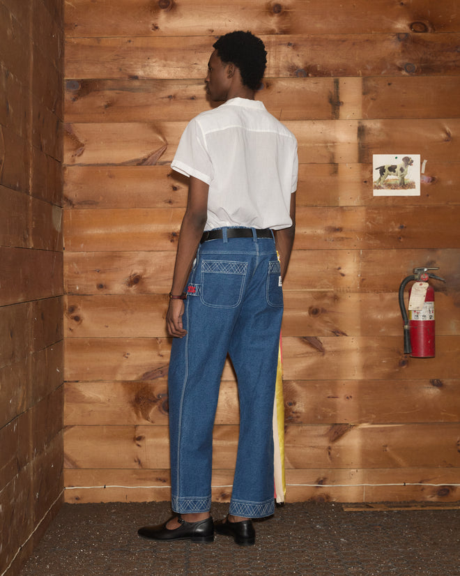 A person in a white shirt and blue jeans stands facing a wooden wall with a fire extinguisher and a dog photo, showcasing the Embroidered Denim Knolly Brook Trousers in a relaxed setting.