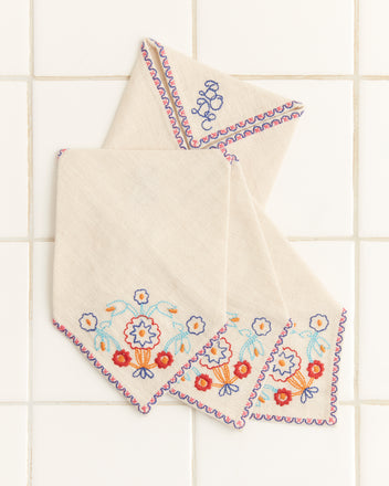 A set of four exquisite linen Garden Party Cocktail Napkins showcases 1950s-inspired floral embroidery and abstract designs, elegantly displayed on a tiled surface.