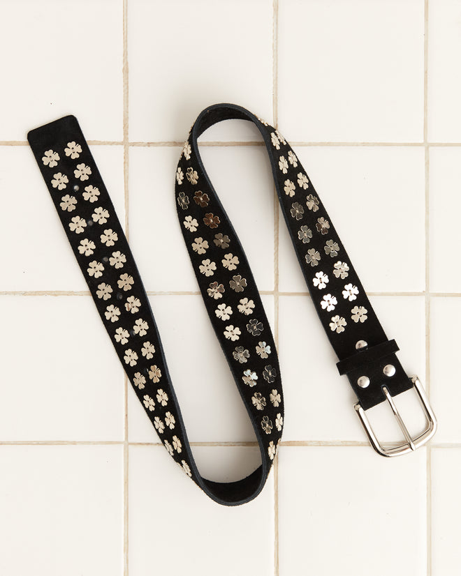 The Clover Charm Belt - Black, decorated with vintage clover charms and silver flower studs, is placed on a tiled white floor.