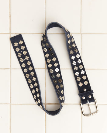 The Clover Charm Belt in dark navy, decorated with silver flower studs and vintage clover charms, rests elegantly on a tiled surface.