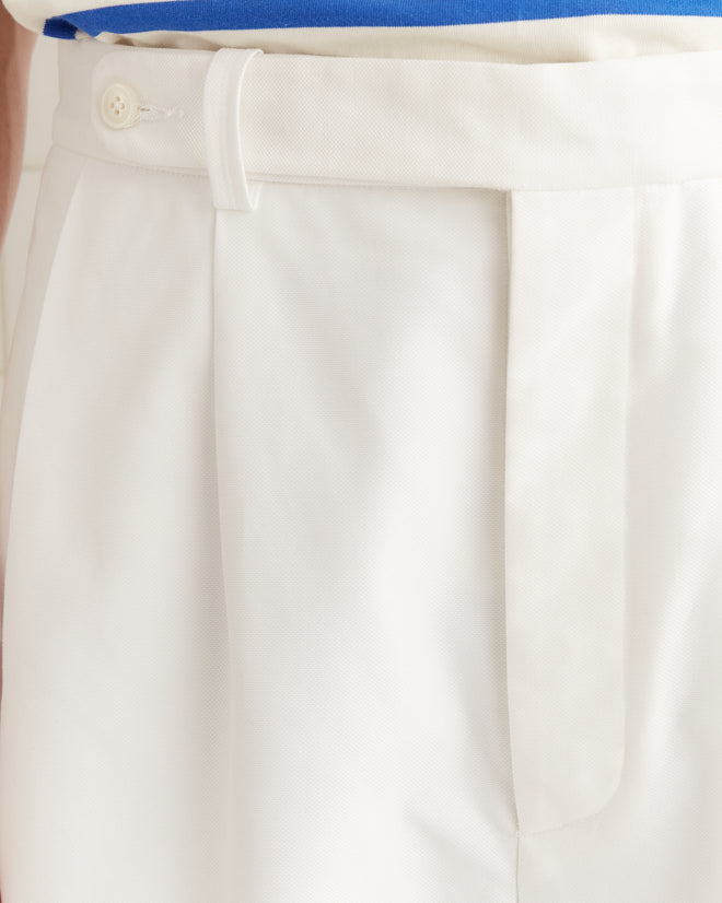 Close-up of a white pleated waistband in piqué cotton with a button and belt loop, paired with a tucked-in blue shirt. The Piqué McNab Trousers provide a modern twist on midcentury style.