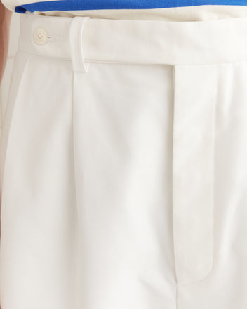Close-up of a white pleated waistband in piqué cotton with a button and belt loop, paired with a tucked-in blue shirt. The Piqué McNab Trousers provide a modern twist on midcentury style.