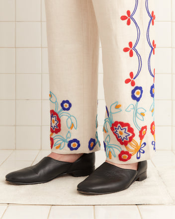 A person in Beaded Garden Party Trousers, adorned with colorful floral patterns, paired with black shoes stands on a white-tiled floor.