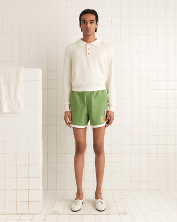 A person in a white sweater and Euro Gym Shorts, embroidered in green, stands in a tiled bathroom near a shower.