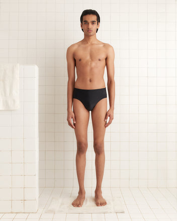 A person stands in a tiled room wearing Swimming Briefs - Black, featuring a visible Bode logo, facing forward with arms at their sides.