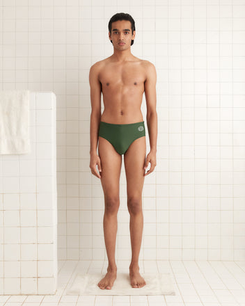 A person in green Swimming Briefs stands in a tiled bath area with crisp white towels, subtly flaunting the Bode logo.