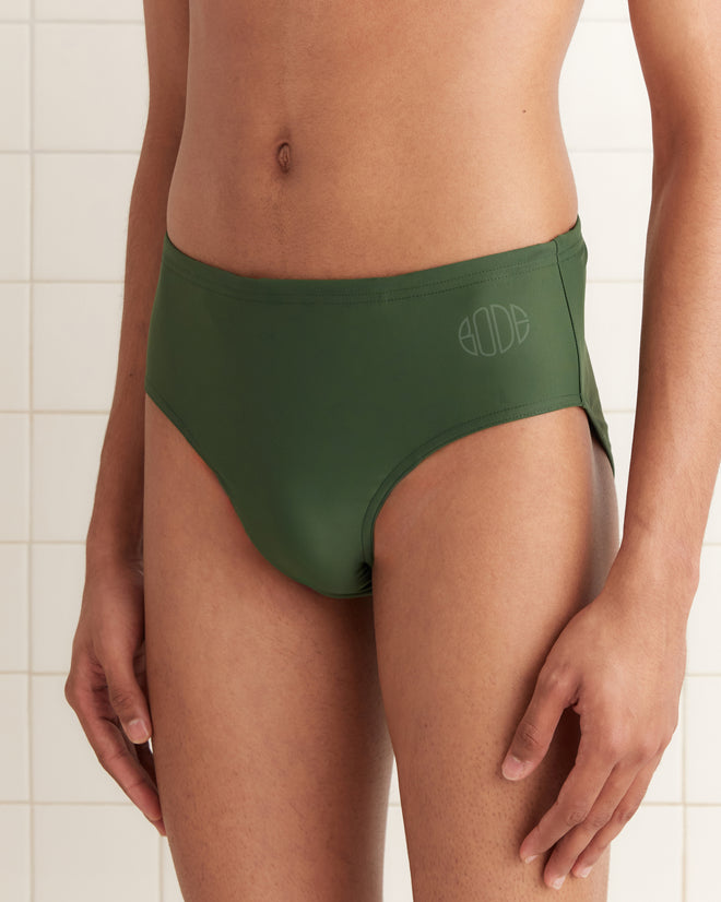 Model wearing Swimming Briefs - Green, made from recycled polyester with a subtle logo detail, standing against a tiled wall.
