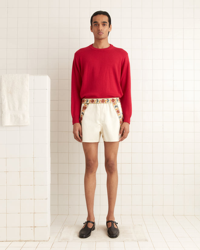 A person in a Juniper Sweater - Pink/Red and white shorts stands in a white-tiled room, exuding classic charm.