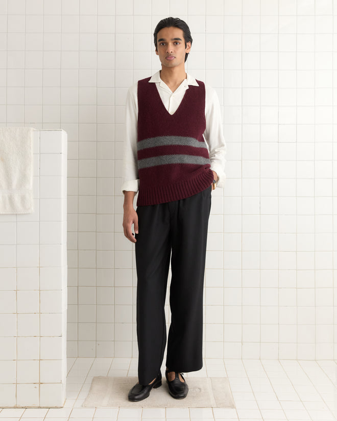 A person in a white shirt, Collegiate Vest from the 1950s, and black pants stands against a tiled wall with a neutral expression.