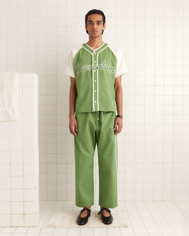 In a green and white outfit, resembling a 1940s reproduction baseball uniform, a person stands against a tiled wall wearing an "Outfield Baseball Shirt.