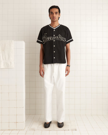 A person in a handmade costume, wearing a Baseball Shirt - Black with star accents and paired with white pants, stands in a tiled room. A towel hanging on the left wall completes the serene setting.