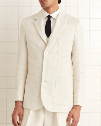 A person confidently stands against a tiled wall wearing the Nelson Single-Breasted Suit Jacket in ivory taupe graph check, reminiscent of 1950s-1960s suiting. The 100% linen jacket pairs perfectly with a classic black tie for a timeless and sophisticated look.