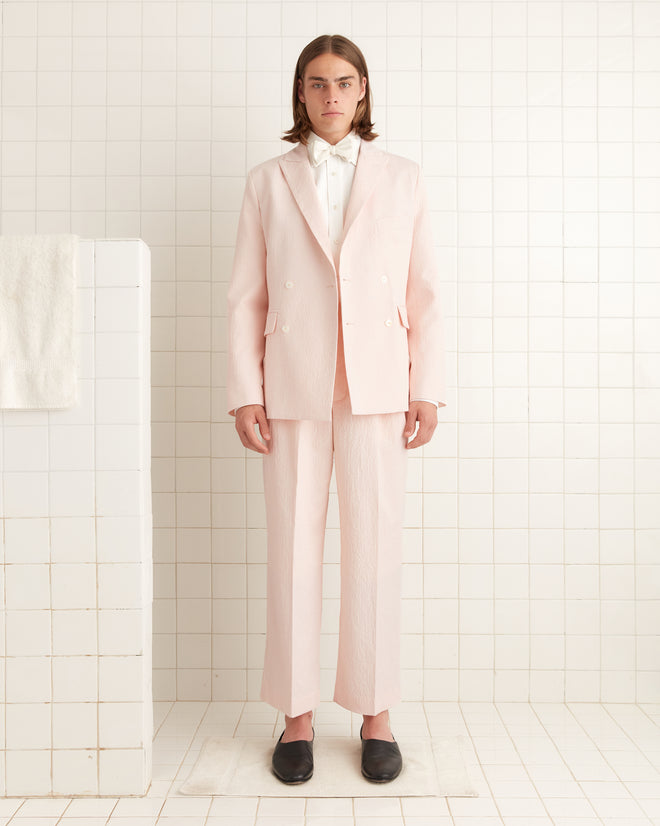 A person wearing a Club Stripe Double-Breasted Suit Jacket in pink/white stands in a tiled room, with a towel on a divider and wearing black shoes.