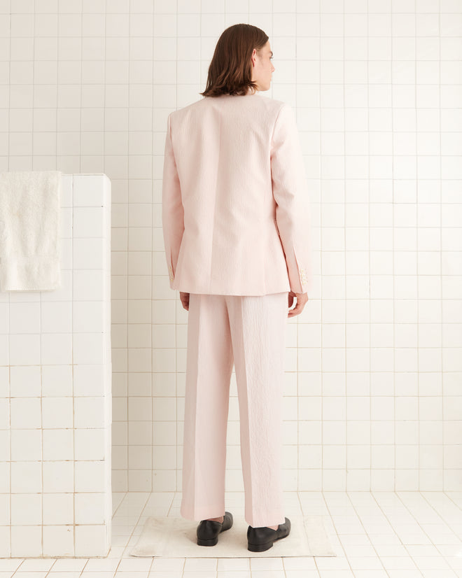 In a tiled room, a person stands facing away, wearing the Club Stripe Double-Breasted Suit Jacket in Pink/White that exudes timeless elegance.