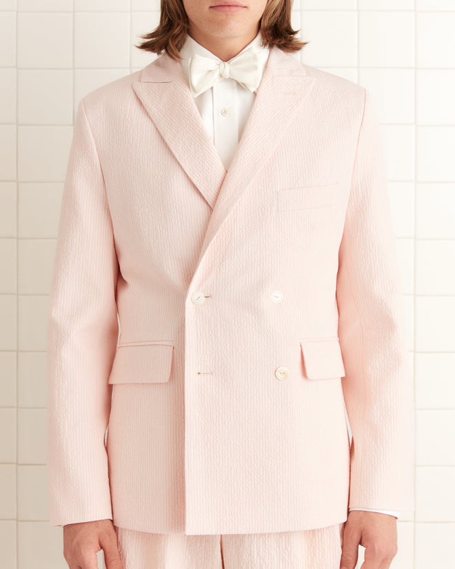 A person confidently stands against a tiled background wearing a Club Stripe Double-Breasted Suit Jacket in Pink/White with a white bow tie.
