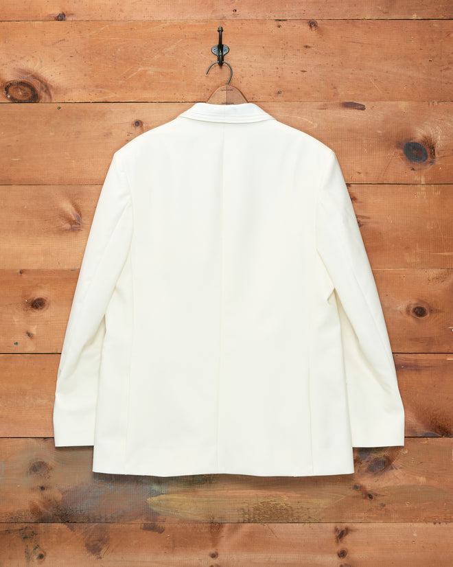 The Meringue Shawl Collar Tuxedo Jacket, crafted from ivory wool with a sleek shawl collar, hangs on a hook against a wooden wall and is displayed from the back.