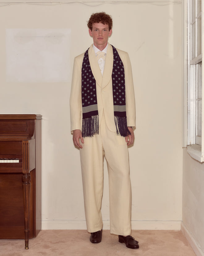 A man in a cream suit and the Foyer Diamond Scarf stands indoors beside a piano and a window.