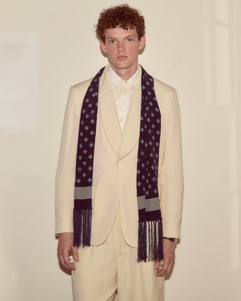 A man in a cream suit and the Foyer Diamond Scarf stands against a plain, light-colored wall, looking at the camera.