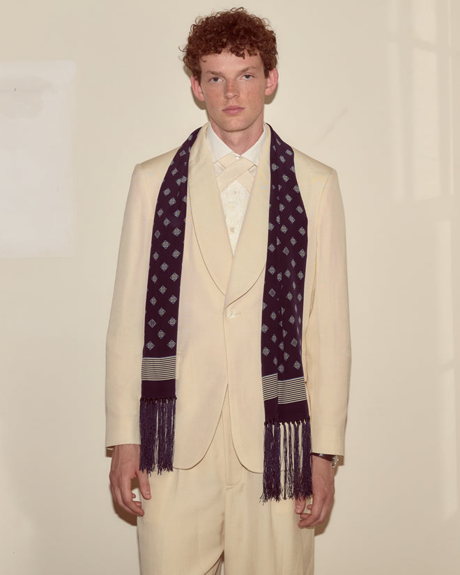 A man in a cream suit and the Foyer Diamond Scarf stands against a plain, light-colored wall, looking at the camera.