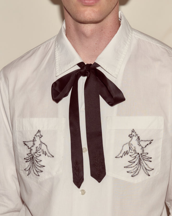 A man wears a white shirt with bird embroidery and wheat motif pockets, paired with the Meadow Soft Tie in black.
