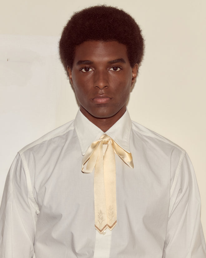 A person wearing a white shirt and the Meadow Soft Tie - Cream, paired with delicate embroidered accessories, stands against a plain beige background.