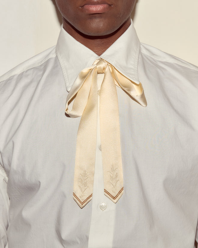 A person wears a white shirt with the Meadow Soft Tie - Cream, a silk ribbon tied in a bow at the collar for an elegant touch.
