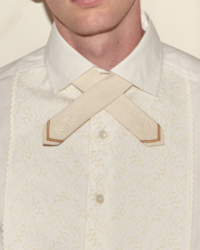 A person wears a white embroidered shirt paired with the Meadow Cross Tie in cream.