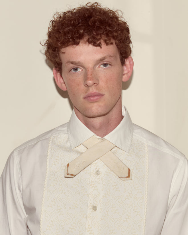 Young person with curly red hair wears a white shirt paired with the Meadow Cross Tie in cream, looking at the camera.