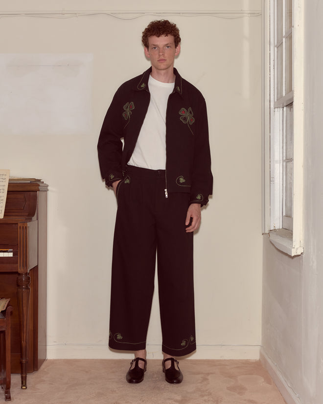 A man stands by a piano and window wearing Lucky Draw Trousers—black, hand-embroidered cotton pants—paired with a white t-shirt and an embroidered suit.