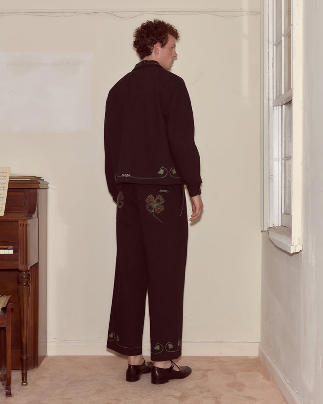 Wearing Lucky Draw Trousers, a person in black hand-embroidered cotton stands by a window in a matching jacket, facing away from the camera.