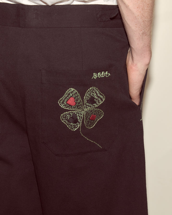 Wearing Lucky Draw Trousers—dark pants featuring a hand-embroidered green clover and 