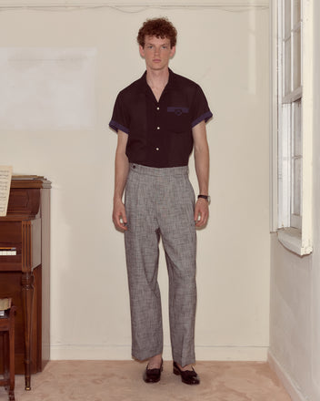 A man stands indoors by a piano, wearing a black shirt, classic suiting with Static Weave McNab Trousers, and black shoes.