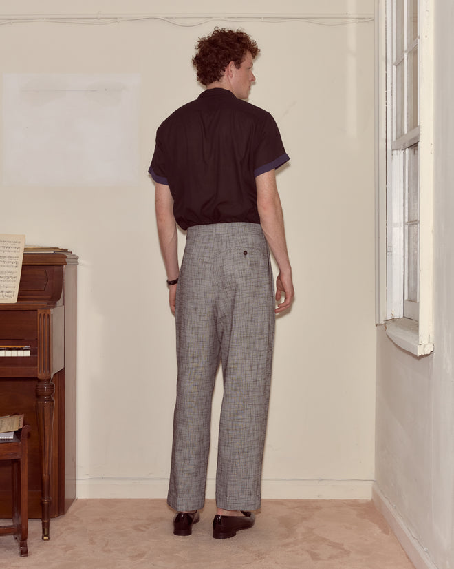 A person stands indoors near a piano and window, wearing a black shirt and Static Weave McNab Trousers, facing the wall.