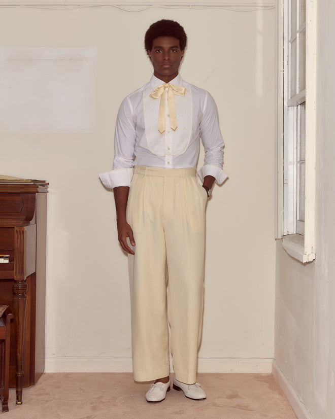 A person stands indoors wearing a white shirt with a bow, Slub Weave Trousers in cream viscose linen, and white shoes, showcasing classic suiting.