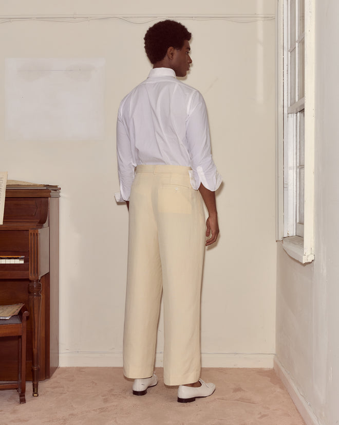 A person in a white shirt and Slub Weave Trousers stands facing a corner in a softly lit room with a piano.
