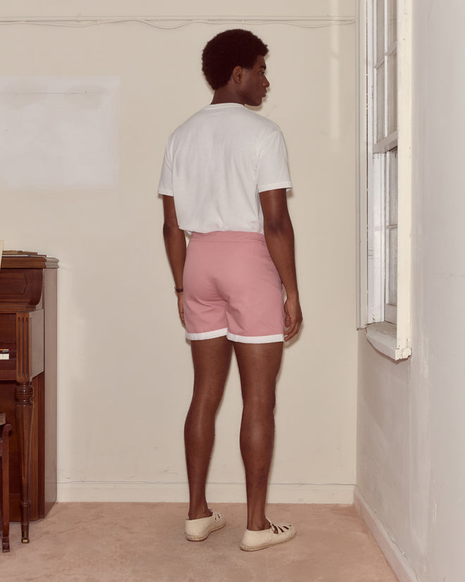 A man in a 100% cotton white shirt and Heart Deco Boxers stands facing a wall in a sparsely furnished room.