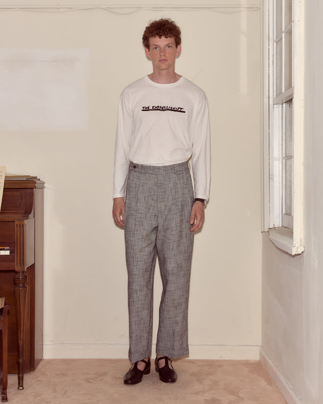 A man stands indoors wearing a white Script Tee and gray plaid trousers, next to a piano.