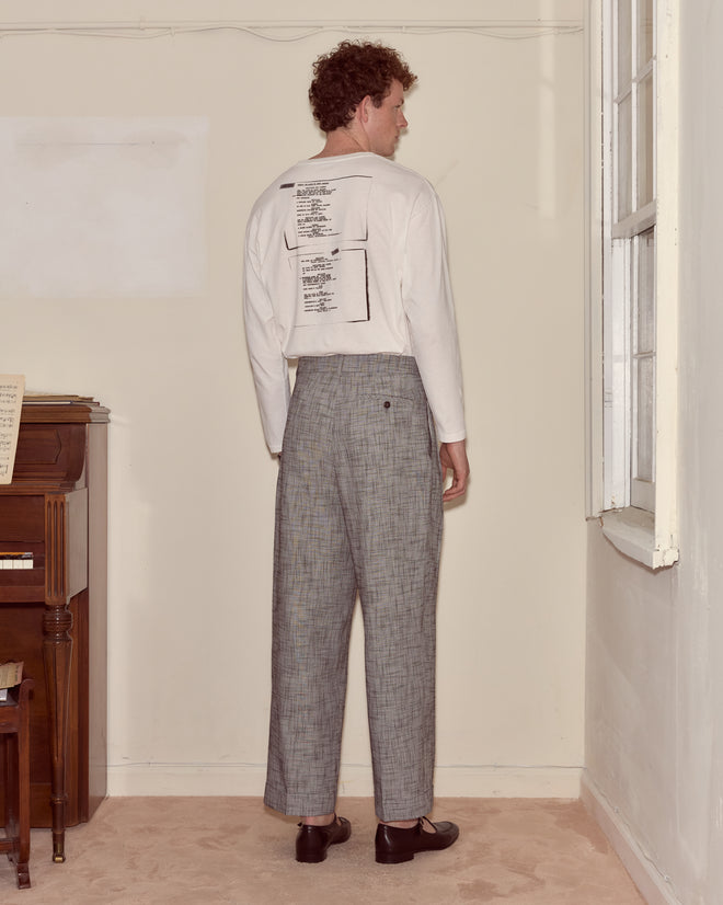 A man stands indoors facing away, wearing a white Script Tee with text and gray plaid pants.