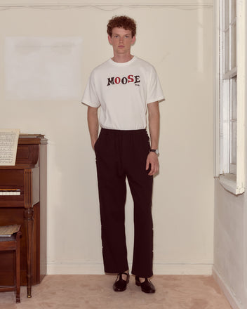 Indoors, a person stands beside a piano wearing a white Moose Tee, black pants, and black shoes.