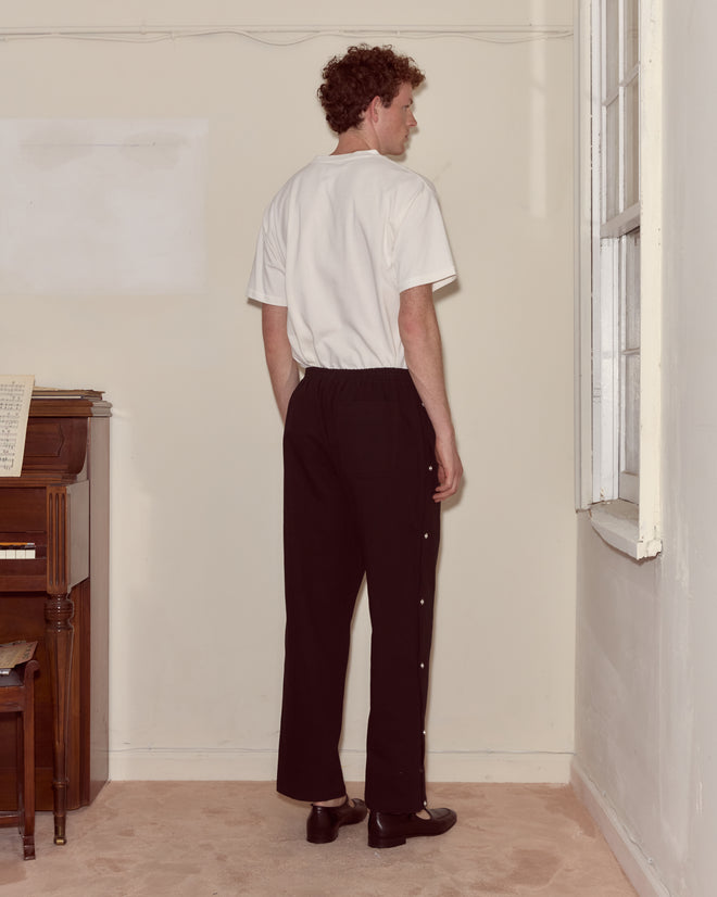 A man with curly hair stands indoors near a window and piano, facing away while wearing a white Moose Tee and black pants.