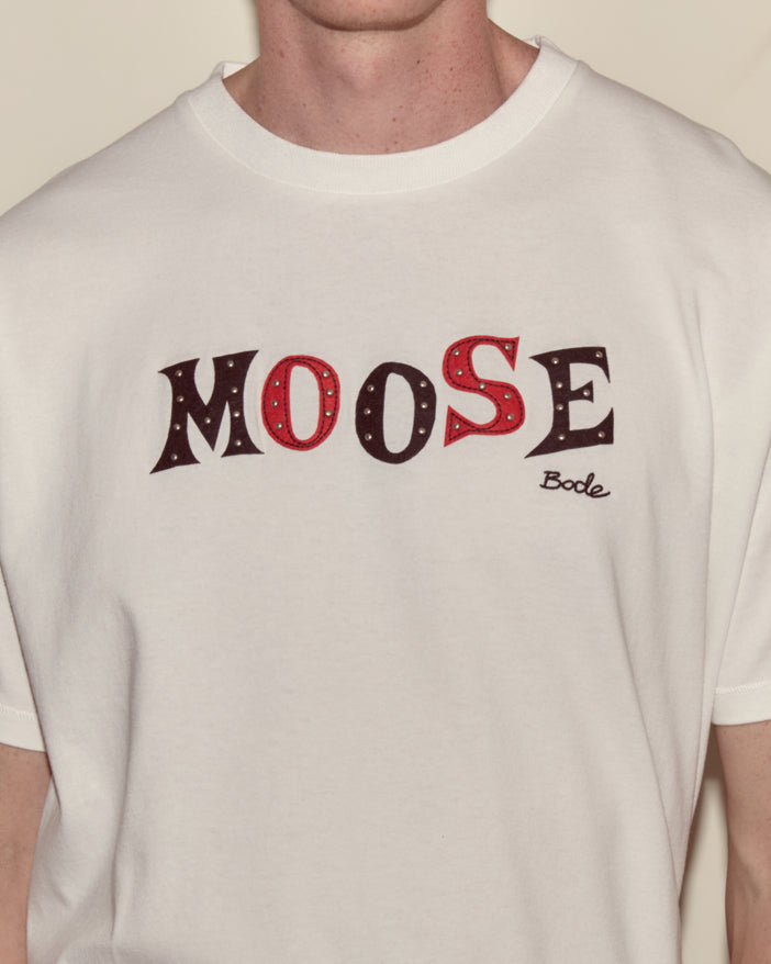 Detail of a men's t-shirt with the name "Moose" appliquéd in red and black felt with silver studs and an embroidered Bode logo.